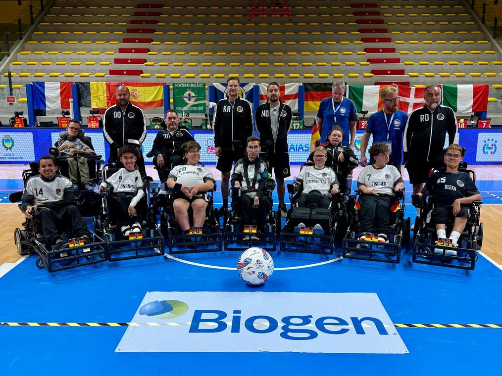 Auf der Fotografie ist die deutsche Nationalmannschaft im E-Rollstuhl Fußball in der Halle in Lignano Sabbiadoro zu sehen. Die Spieler befinden sich mitsamt ihren Rollstühlenstühlen in der ersten Reihe, die Trainer reihen sich dahinter ein. Vor allen liegt ein Ball in der Mitte des Bildes. Foto: Agentur Frontcourt