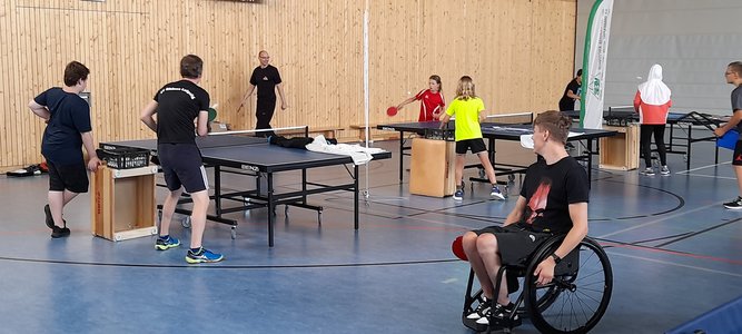 Menschen spielen Tischtennis in einer Sporthalle. Im Vordergrund ist ein Rollstuhlfahrer sichtbar.