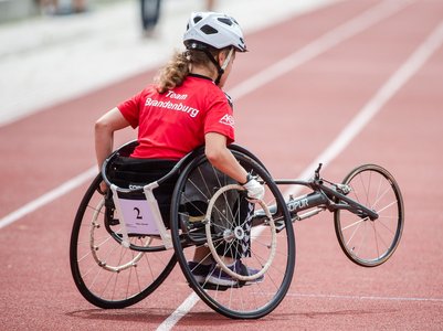 Eine Sportlerin in einem Rennrollstuhl auf einer roten Laufbahn. Sie trägt ein rotes T-Shirt mit der Aufschrift "Team Brandenburg" auf dem Rücken, einen weißen Helm und schwarze Handschuhe. Der Rennrollstuhl hat zwei große Hinterräder und ein kleines Vorderrad, die Sportlerin sitzt nach vorne geneigt und bewegt sich mit den Händen an den Hinterrädern vorwärts.