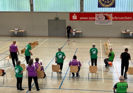 Auf dem Bild sieht man die Spielerinnen und Spieler des SV Rhinos Leipzig (in lila Trikots) im Spiel gegen die Mannschaft aus Köthen (grüne Trikots). Am Ende des Spielfeldes befindet sich ein Linienrichter und beobachtet die Spielsituation genau. Alle Teilnehmenden warten gespannt auf das Ergebnis der nächsten Aktion.