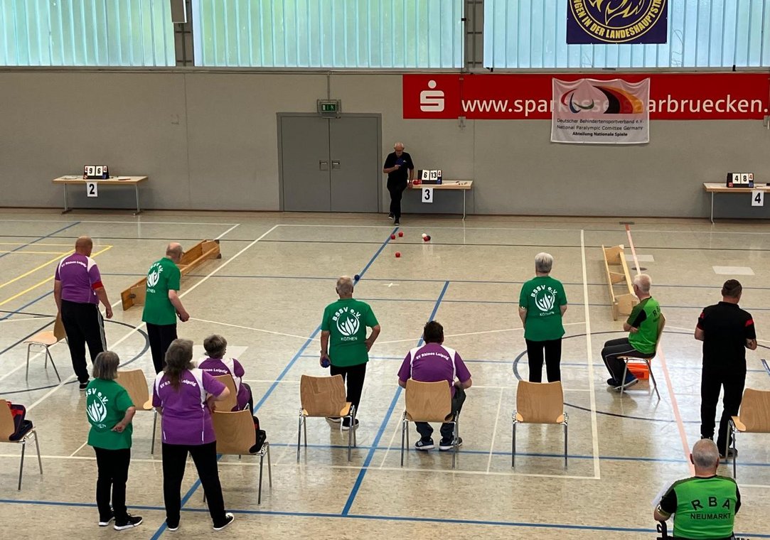 Auf dem Bild sieht man die Spielerinnen und Spieler des SV Rhinos Leipzig (in lila Trikots) im Spiel gegen die Mannschaft aus Köthen (grüne Trikots). Am Ende des Spielfeldes befindet sich ein Linienrichter und beobachtet die Spielsituation genau. Alle Teilnehmenden warten gespannt auf das Ergebnis der nächsten Aktion.