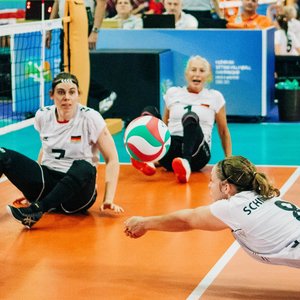 Bild der Deutschen Sitzvolleyball-Nationalmannschaft während eines Spiels. In der Bildmitte hechtet eine Spielerin zum Balll