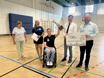 Pokal wird Rollstuhlfahrer Josco Wilke übergeben. Wilke streckt den Pokal jubelnd mit der linken Hand in die Luft. Ein Scheck mit 1000€ wird vom Sportbürgermeister der Stadt Leipzig festgehalten. Foto: Kristin Jorks